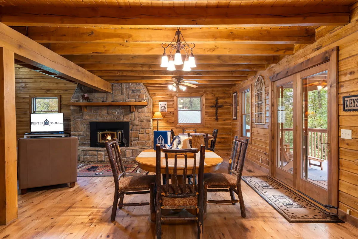 This image beautifully captures the main gathering space, showcasing a large wooden dining table, a cozy wood-burning fireplace, exposed wooden beams, and expansive windows framing views of the forest. The atmosphere is warm and inviting, with seating and layout that encourage family gatherings, meals, and relaxation by the fire.