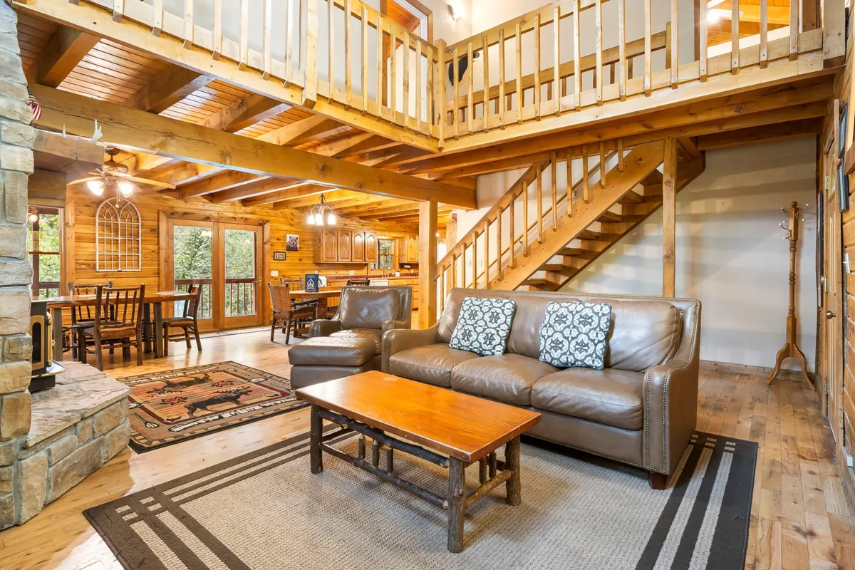 The image showcases a spacious log cabin interior with a towering stone fireplace, a sweeping wooden staircase, plush seating, and a large dining area. The warm wood tones, cozy furnishings, and inviting decor create a welcoming space perfect for family gatherings and relaxation.