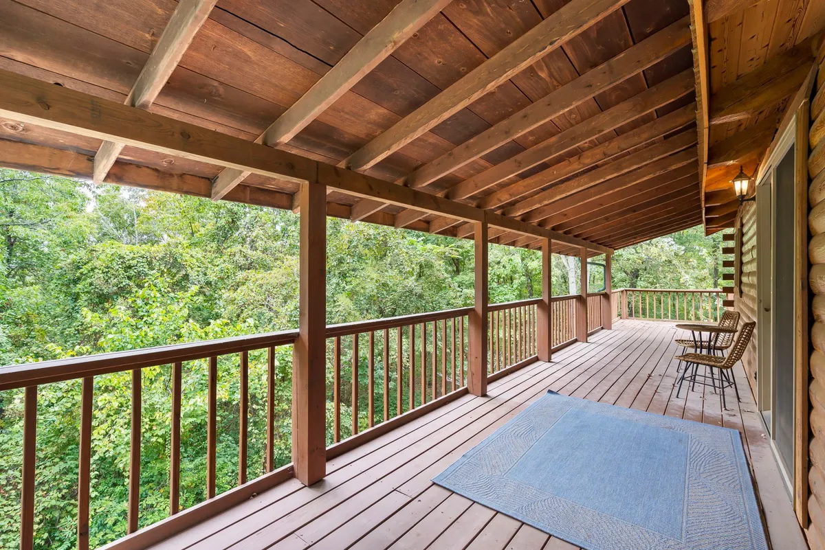 A relaxed outdoor sitting area on a deck, with wooden railings, comfortable chairs, a blue area rug, and lush greenery in the background. The setup invites guests to enjoy nature views, read, or simply soak in the calm, evoking both summer evenings and year-round enjoyment of the outdoors.