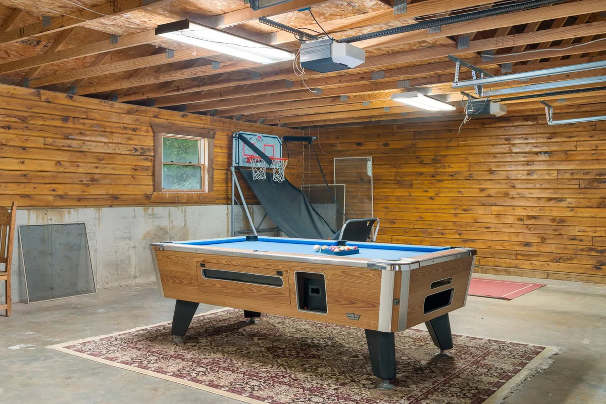 This image shows a converted garage game room featuring a pool table, basketball hoop, and wooden walls. The space is well-lit, emphasizing its playful and relaxed atmosphere, and clearly centers on recreational activities like pool and shooting hoops.