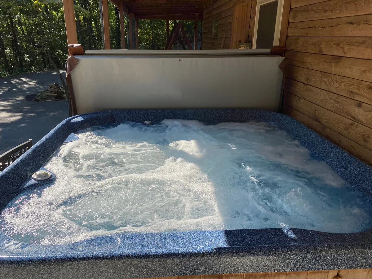 The exterior shot features a wooden cabin with a bubbling hot tub nestled against a backdrop of dense forest. The scene suggests relaxation after a day outside, with ample space for groups to unwind in privacy.