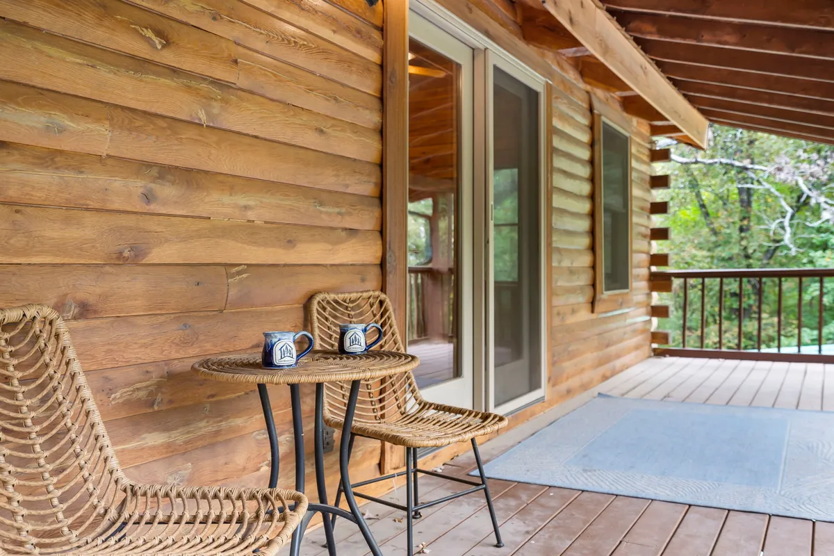 This exterior image features a pair of woven chairs and coffee mugs set against a wooden wall, creating an inviting space for morning coffee or relaxed conversation. The natural surroundings beyond hint at ample private acreage for kids to play and guests to unwind, capturing the tranquil outdoor experience central to the cabin's summer highlights.