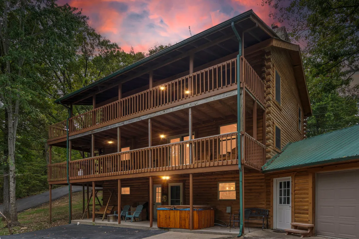 A wide view of the cabin's three levels, showcasing wooden balconies and a private hot tub set against a forested backdrop. Guests are pictured enjoying the hot tub in the foreground and gathering on balconies, hinting at the ample outdoor spaces for groups.