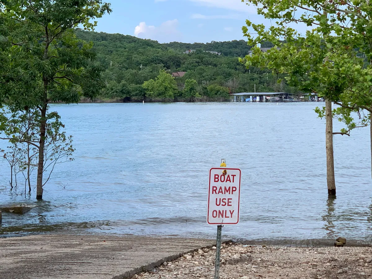 This image captures the calm water near a boat ramp, surrounded by lush trees and natural shoreline. The boat ramp sign is visible, inviting visitors to set off on boating or fishing adventures. The tranquil mood and the accessible water edge perfectly evoke the spirit of lakeside excursions.