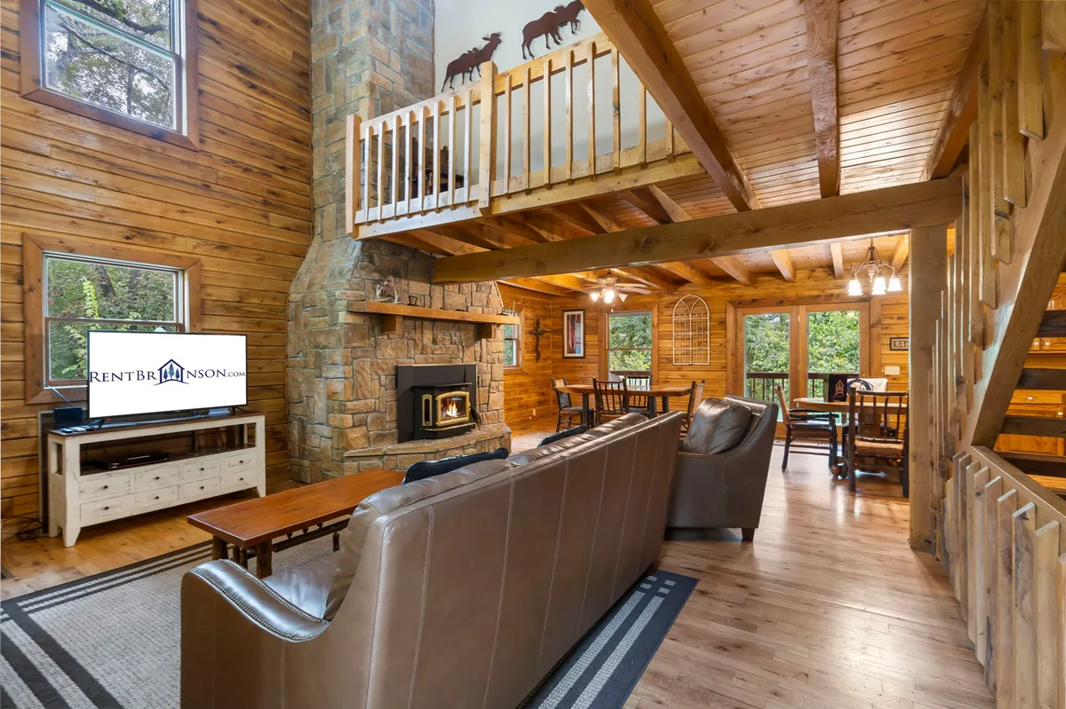 The living area is shown with comfortable sofas arranged around a fireplace and a large television, with the dining space visible in the background. The open design and soft lighting create a welcoming setting for movie nights, shared meals, and relaxed evenings with loved ones.