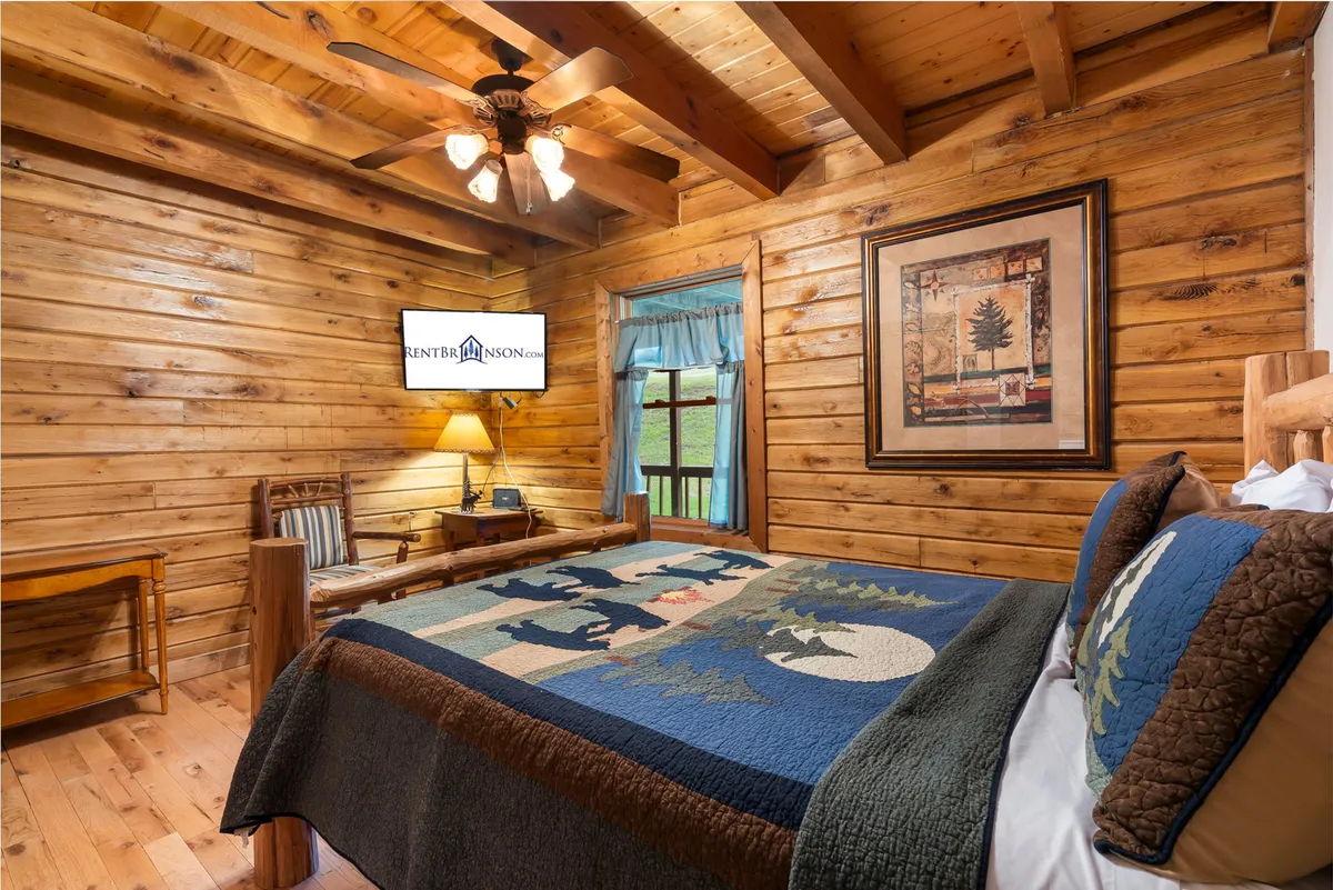 A comfortable bedroom with a colorful quilt on the bed, wooden ceiling beams, a TV mounted on the wall, and rustic décor. The space is both restful and equipped for entertainment.