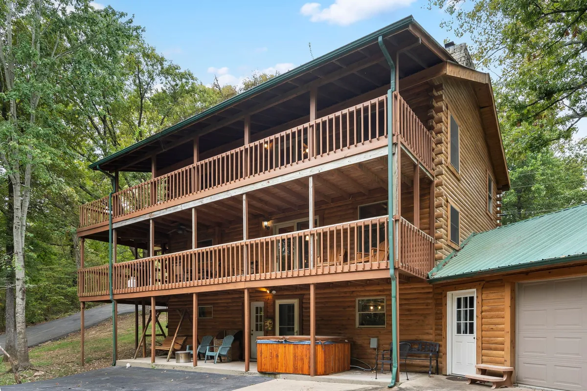 A spacious, multi-level wooden deck nestled in a natural setting, featuring a bubbling hot tub, large windows looking out to the forest, and ample room for outdoor relaxation and gatherings. The deck is surrounded by lush greenery, providing privacy and a tranquil backdrop.