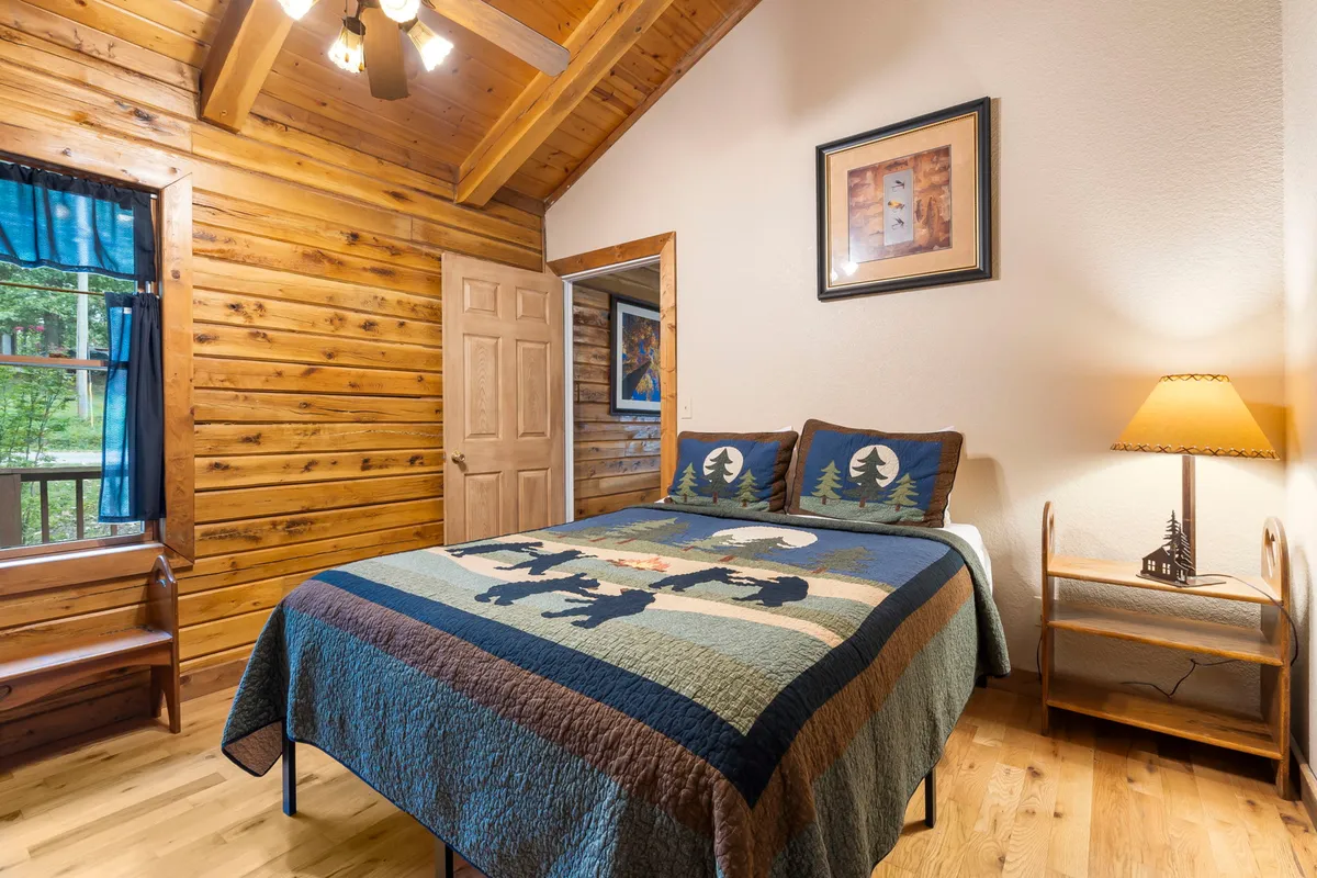 A cozy bedroom with a bed covered in a quilted blanket, wood-paneled walls, a bedside lamp, and framed artwork. The warm lighting and inviting decor create a serene spot for rest and relaxation.
