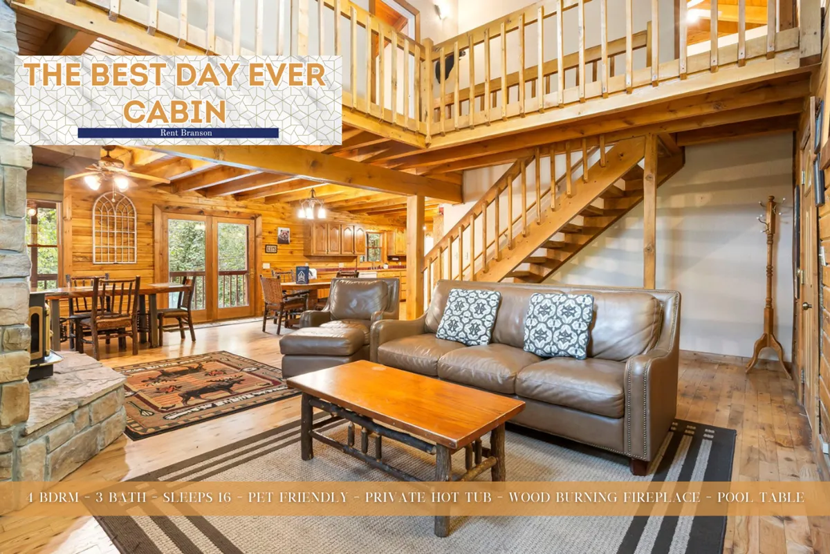 A cozy and spacious interior scene featuring wooden beams, a plush leather sofa, a stone fireplace, a dining table, and a glimpse of stairs leading to other levels. The room is set up for both relaxation and lively gatherings, reflecting the cabin’s versatility.